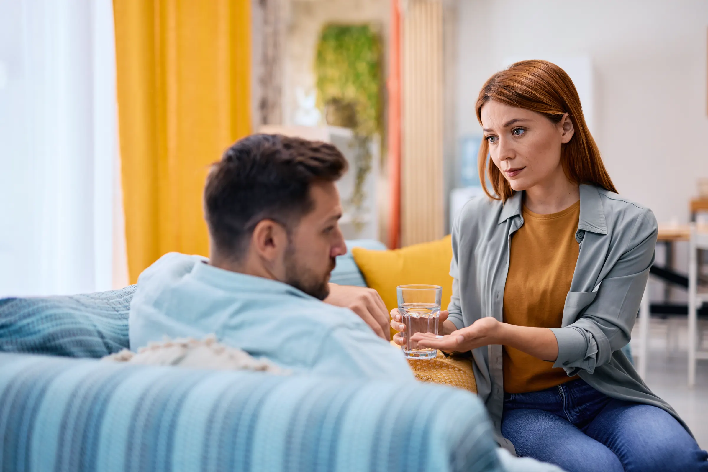 Frau reicht ihrem erkrankten Partner auf dem Sofa ein Glas Wasser