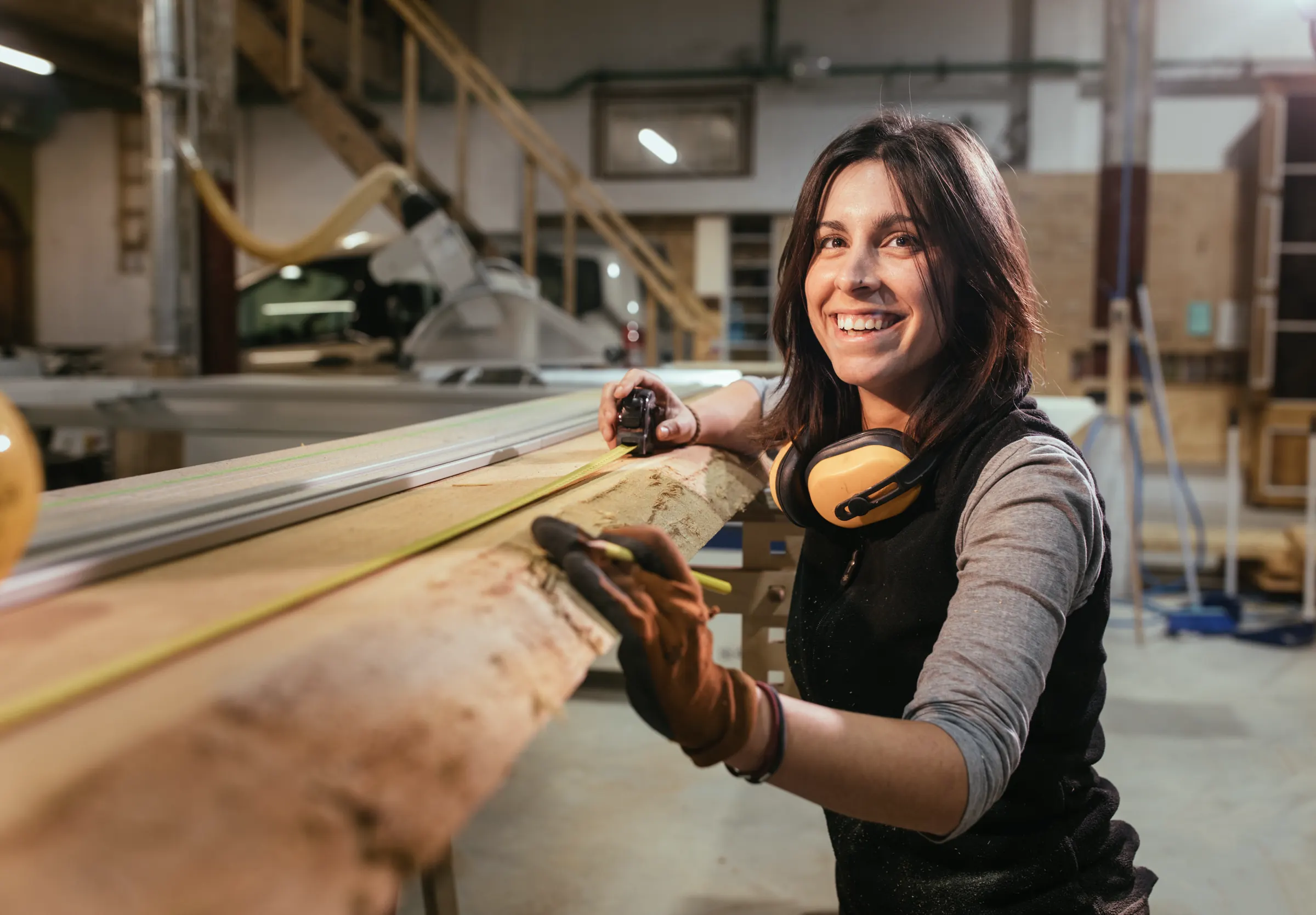 Tischlerin mit Gehörschutz prüft lachend ein Holzbrett in der Werkstatt