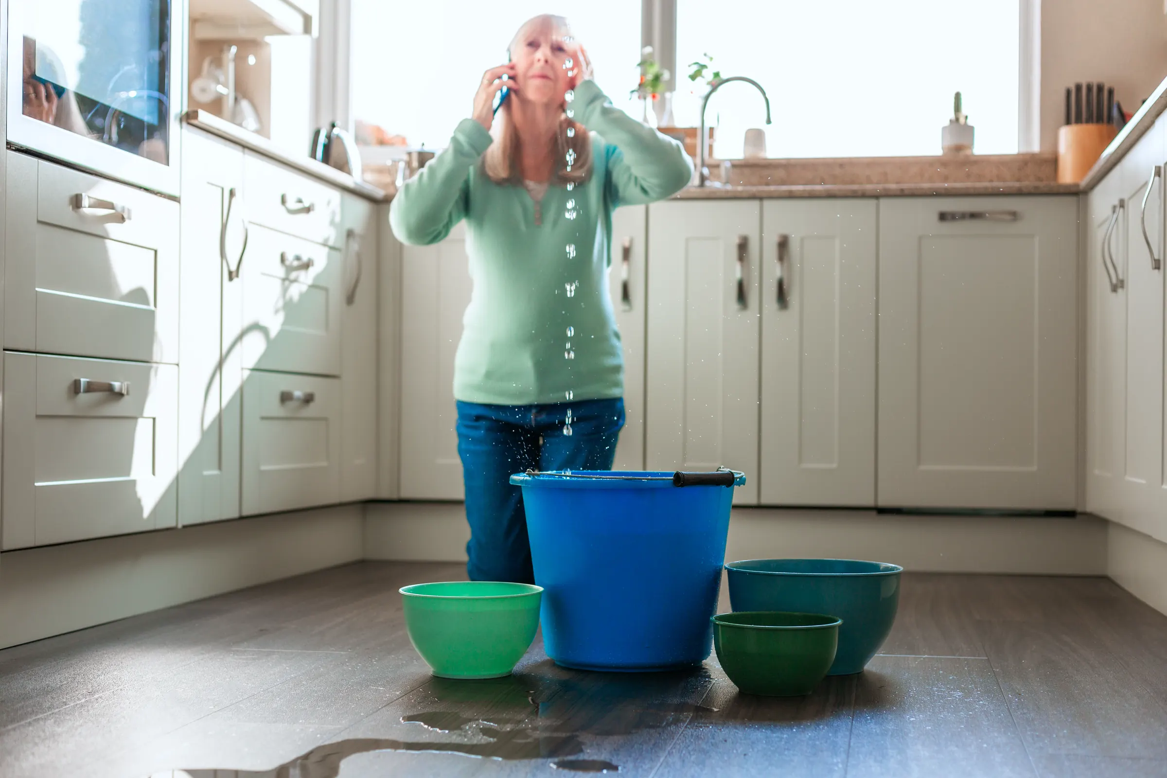 Frau telefoniert in der Küche, vor ihr Eimer unter einem Wasserleck