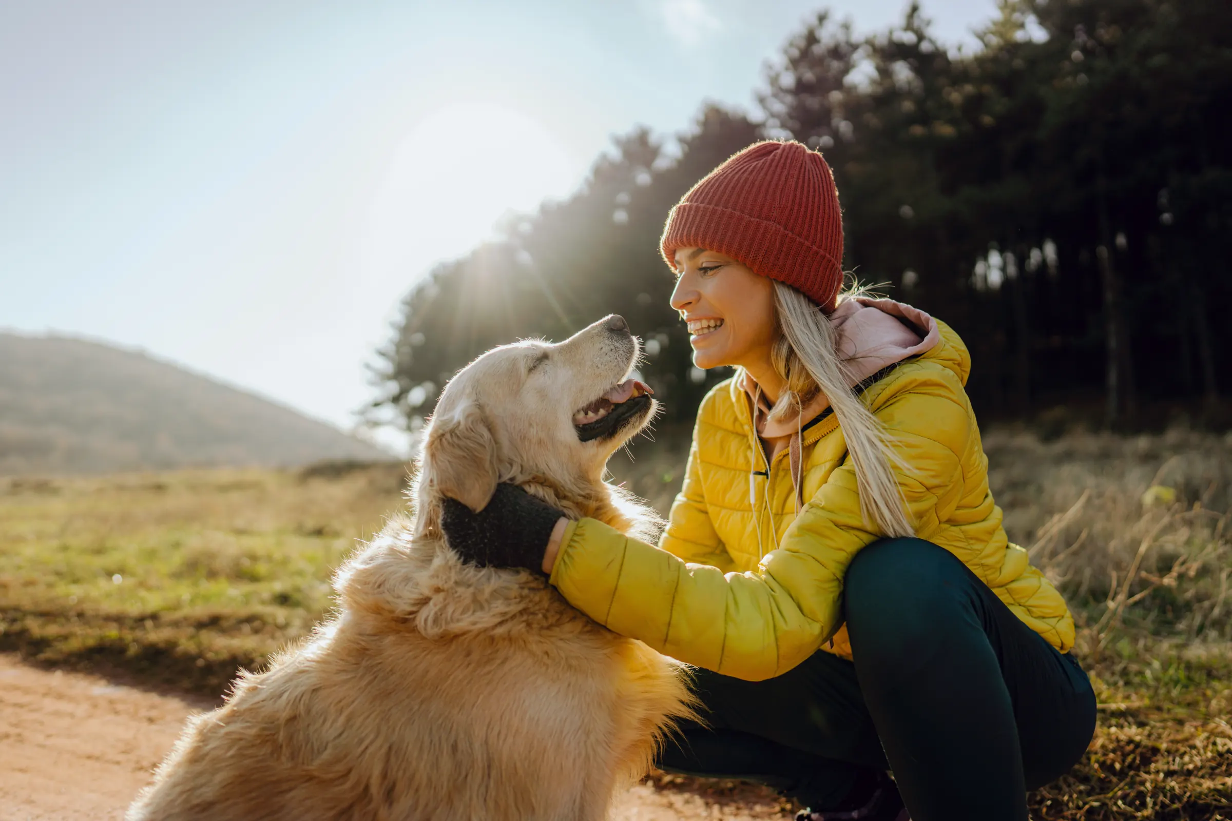 Frau mit Mütze knuddelt draußen im Gegenlicht ihren Golden Retriever