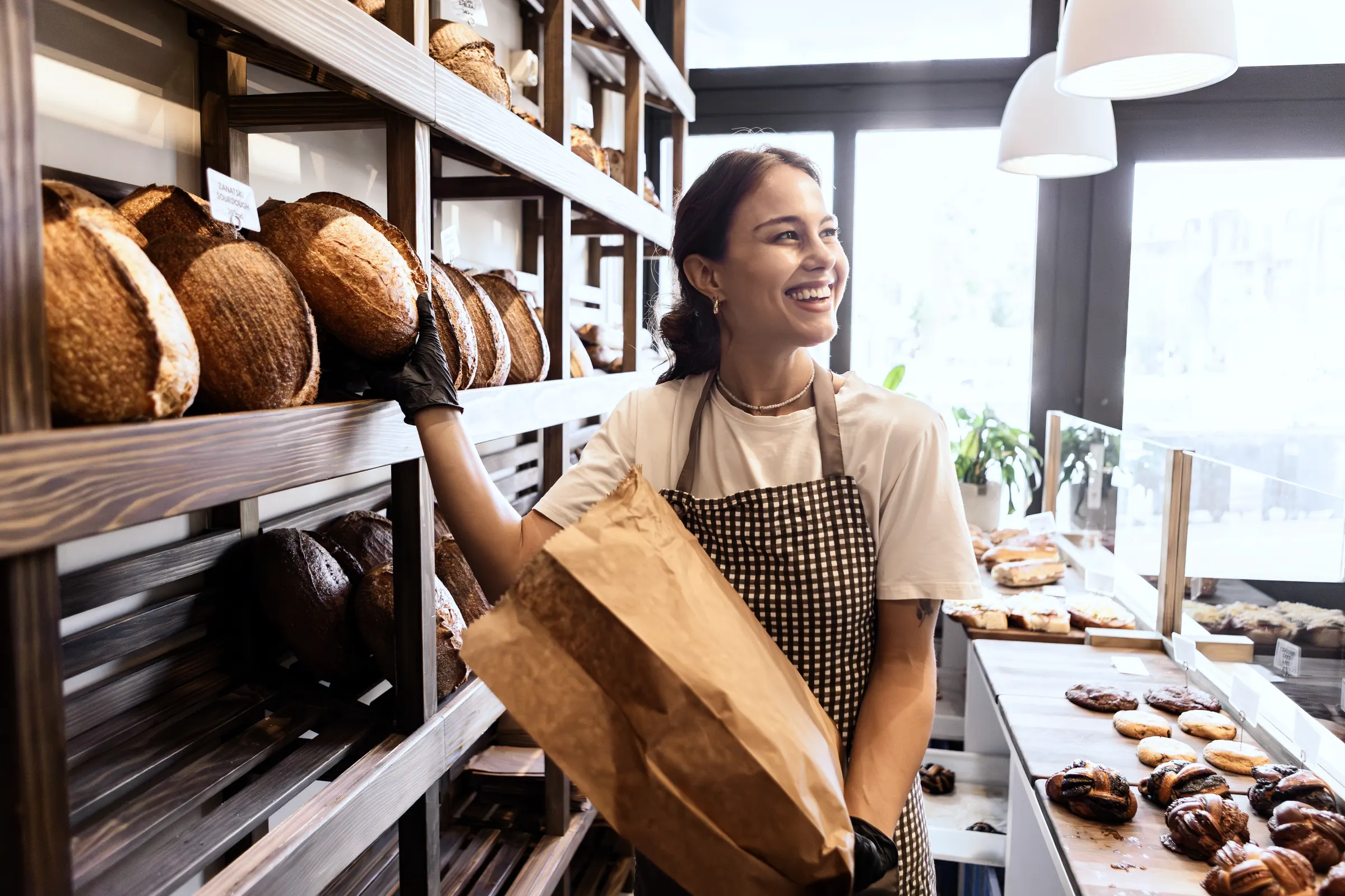 Bäckerin reicht lächelnd frisches Brot in einer Papiertüte über die Theke