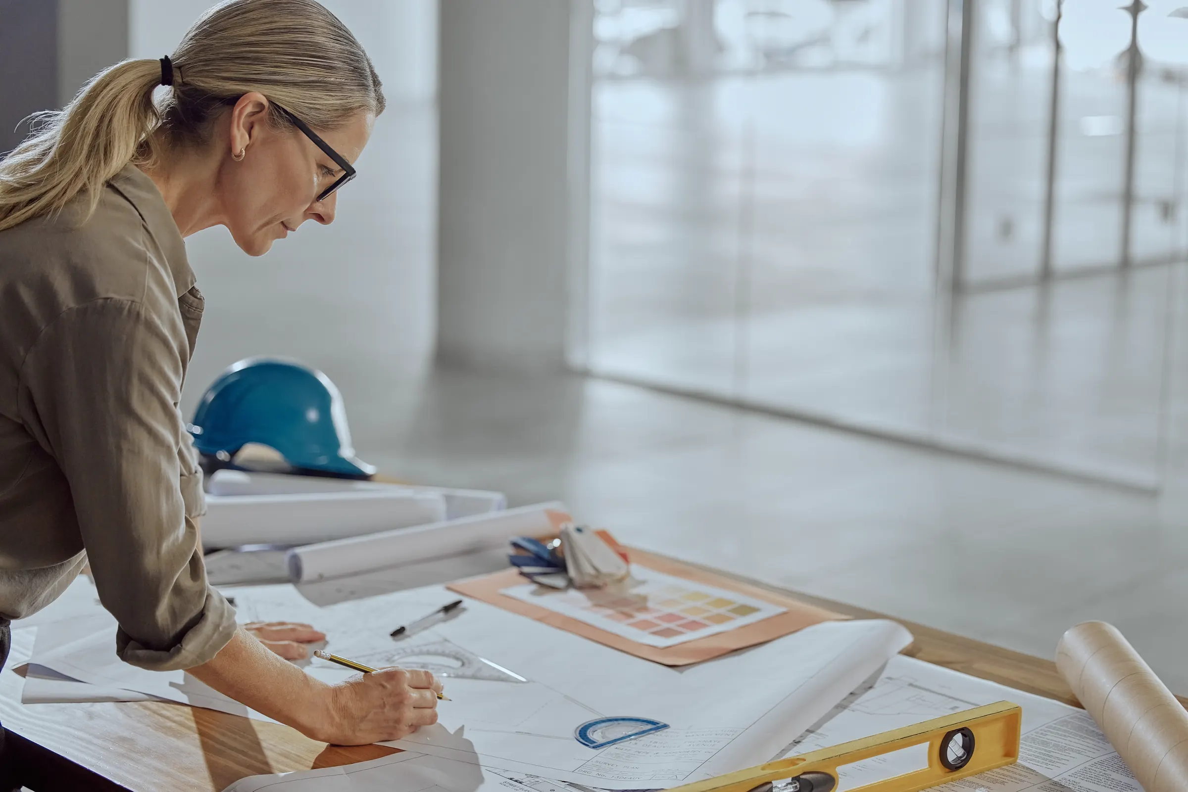 Architektin zeichnet im hellen Büro an Bauplänen, daneben ein Schutzhelm