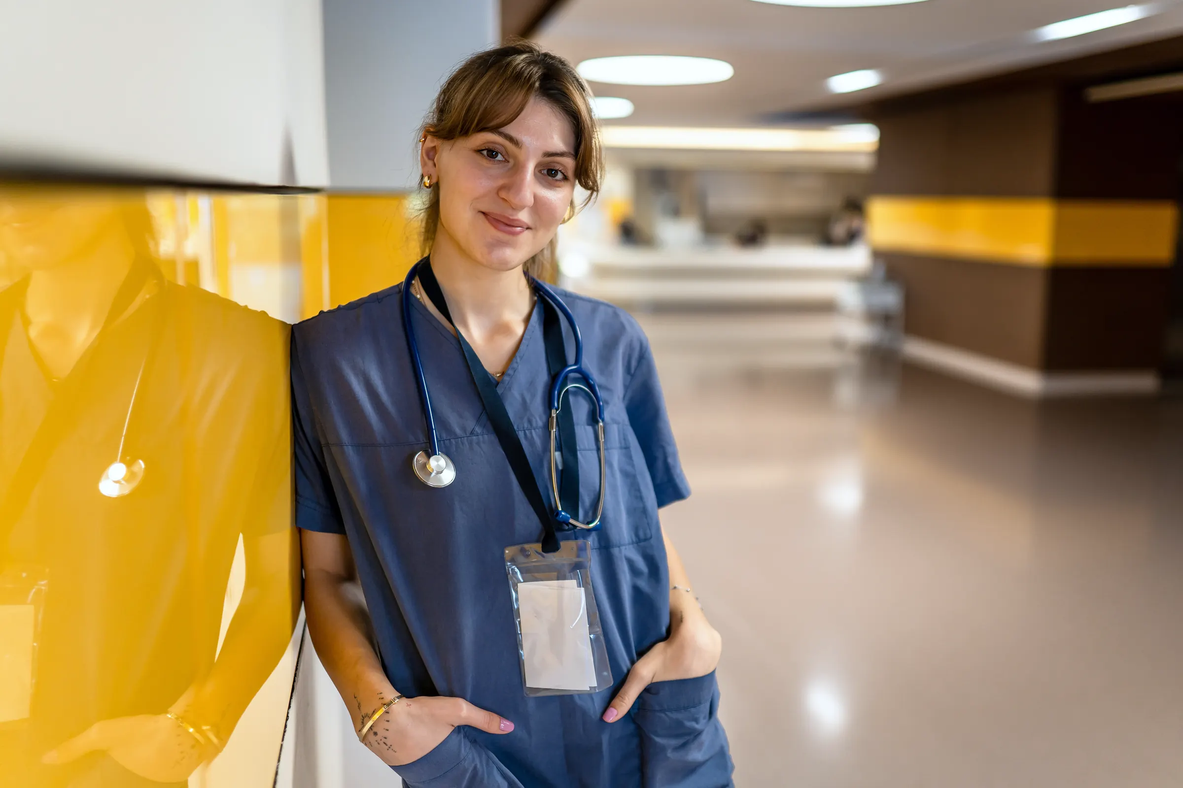 Junge Pflegekraft in Dienstkleidung lehnt lächelnd an der Wand eines Klinikflurs