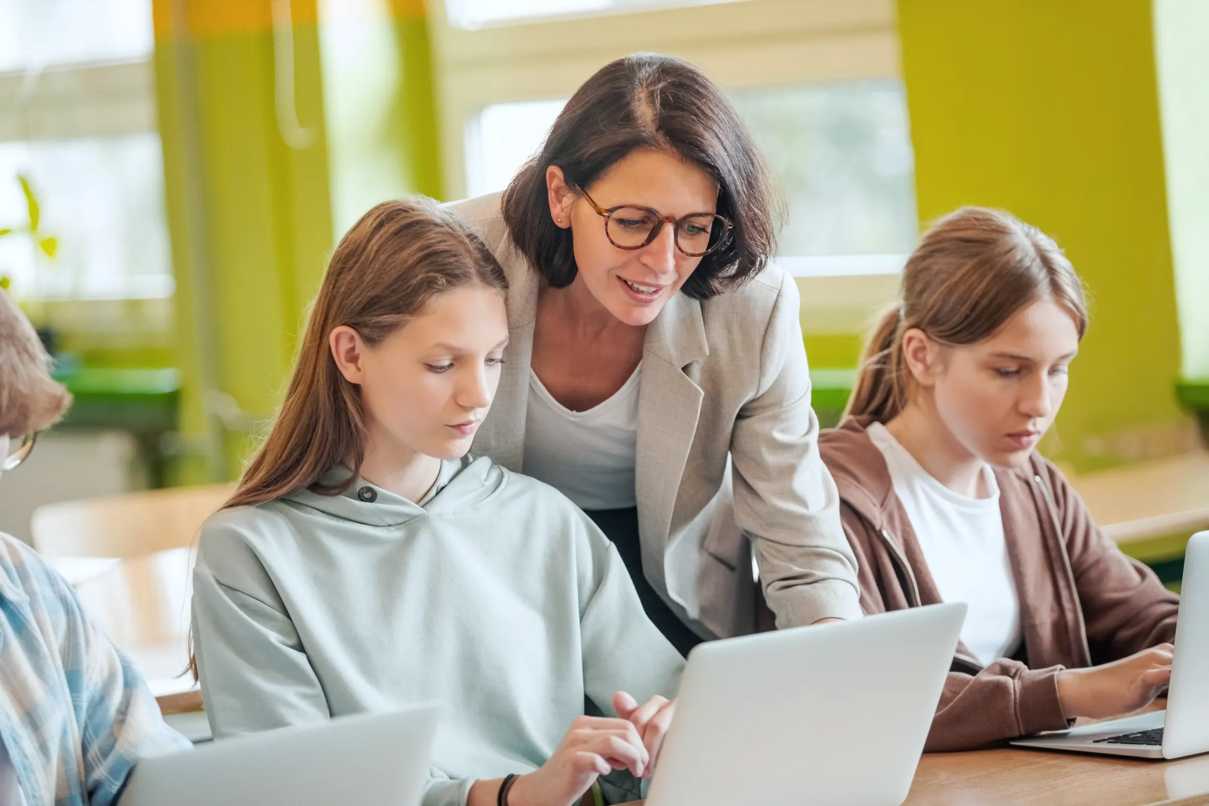Lehrerin hilft zwei Schülerinnen im Klassenzimmer am Laptop