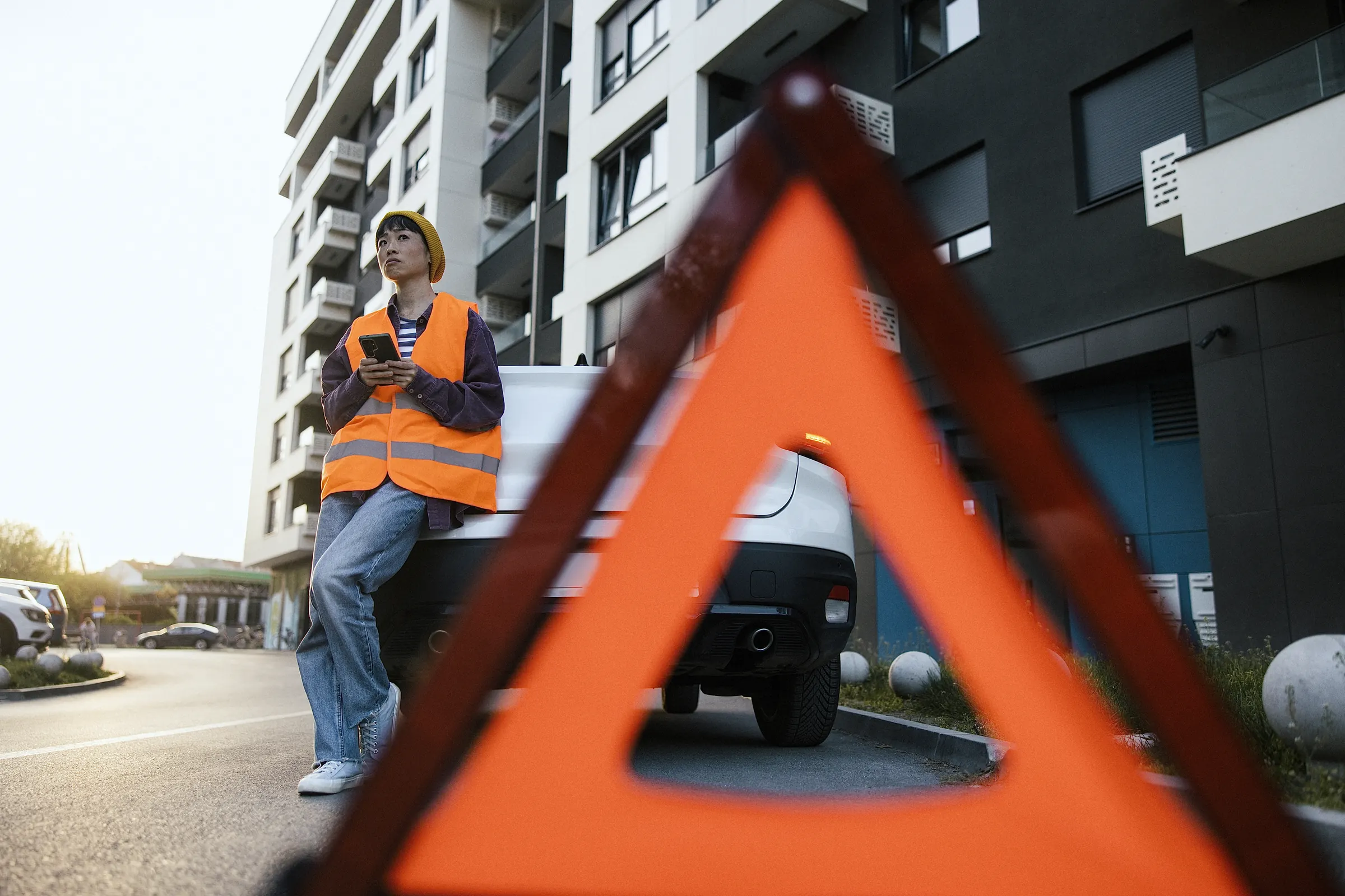 Warndreieck im Vordergrund, dahinter telefoniert eine Frau mit Warnweste an ihrem liegengebliebenen Auto